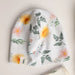 A baby beanie hat with a white background and a colorful floral and fern pattern, featuring yellow flowers, pink blossoms, and green leaves. A small fabric tag on the brim reads 'NBL'.