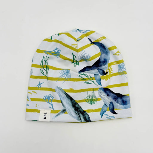 A baby beanie hat featuring a watercolor print of whales and sea plants on a white background with horizontal yellow stripes. A small white tag on the left side displays the brand 'NBL'.