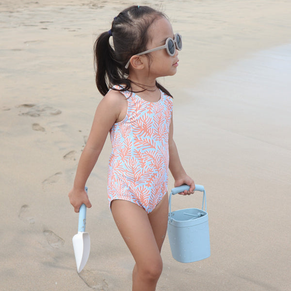 Child in a swimsuit with a leaf pattern on a beach holding a sand bucket and shovel.