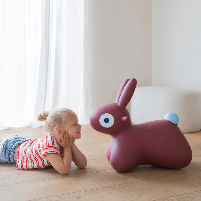 Child lying on the floor next to a pink bunny-shaped toy in a bright room.