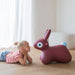 Child lying on the floor next to a pink bunny-shaped toy in a bright room.
