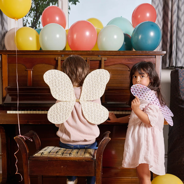 Two children with butterfly wings standing in front of a piano with colorful balloons.