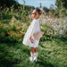 Young girl in a white dress holding a teddy bear in a garden with wildflowers.