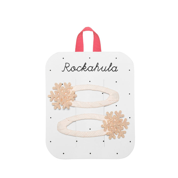 A white display card with the brand name 'Rockahula' printed at the top, holding two cream-colored hair clips decorated with beige glitter snowflake charms.
