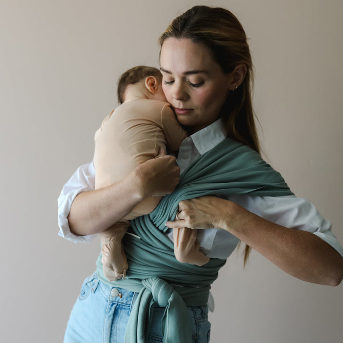Woman holding a baby wrapped in a teal sling against a plain background