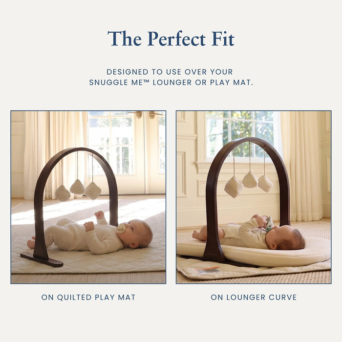 A baby activity gym with an arch-shaped dark wood frame and three hanging fabric toys, shown in two inset photos on a light beige background. Text at the top reads 'The Perfect Fit' and explains how it fits over a Snuggle Me lounger or play mat.