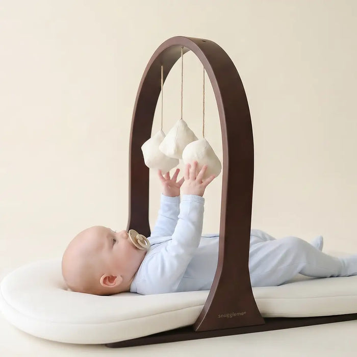 baby reaches for cloth toys attached to a wooden toy bar set against a cream background