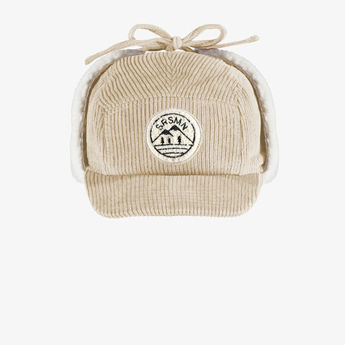 Beige corduroy cap with ear flaps tied up on top of hat on white background