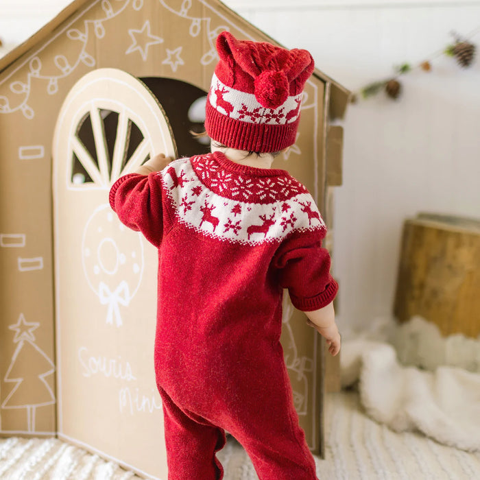 Child wearing a red Christmas-themed outfit standing in front of a wooden toy house.