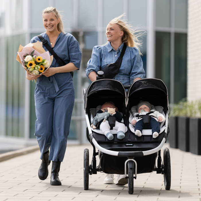 Two women walking with a Thule stroller carrying two children, one holding flowers.