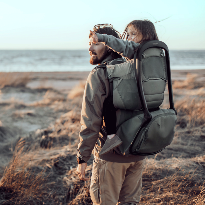 Man carrying a child on his back with a backpack, standing on a beach.