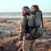 Man carrying a child on his back with a backpack, standing on a beach.