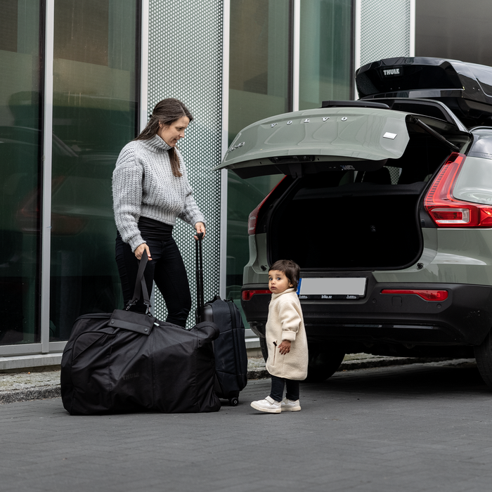 Woman with a child standing next to a car with an open trunk, preparing for a journey with stroller travel bag