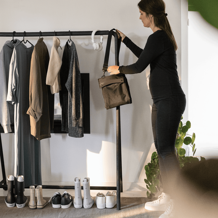 Woman selecting a folded up hanging travel stroller bag from a rack of clothes with shoes below.