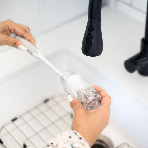 Person using a bottle brush to clean a glass bottle under running water.