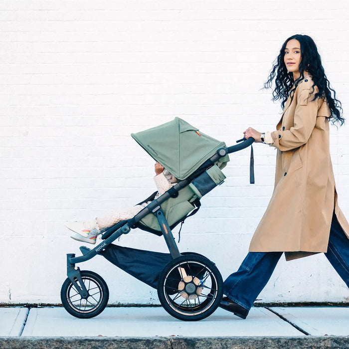 Woman pushing a green stroller with a child inside against a white wall.