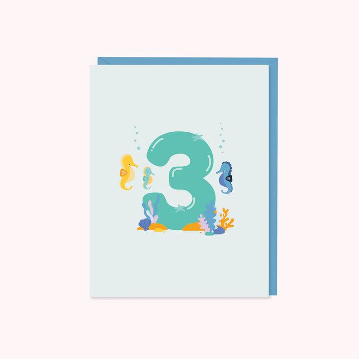 A "3" is featured with 3 seahorses surrounding the shape as if it's on a seabed and a few ocean plants are nearby. Original design by Halifax Paper Hearts