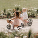 Image is of two children on wooden pink tricycles riding slightly down a hilly bike path with flowers on each side on a sunny day. Helmet are strongly suggested