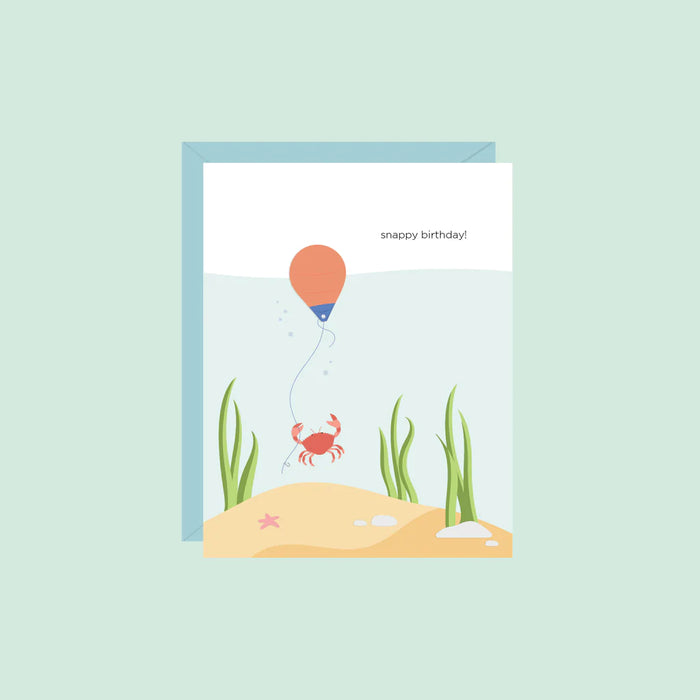 A crab with beach scene behind is holding on to a buoy in the shape of a balloon with string. Caption reads "Snappy Birthday" original design by Halifax Paper Hearts